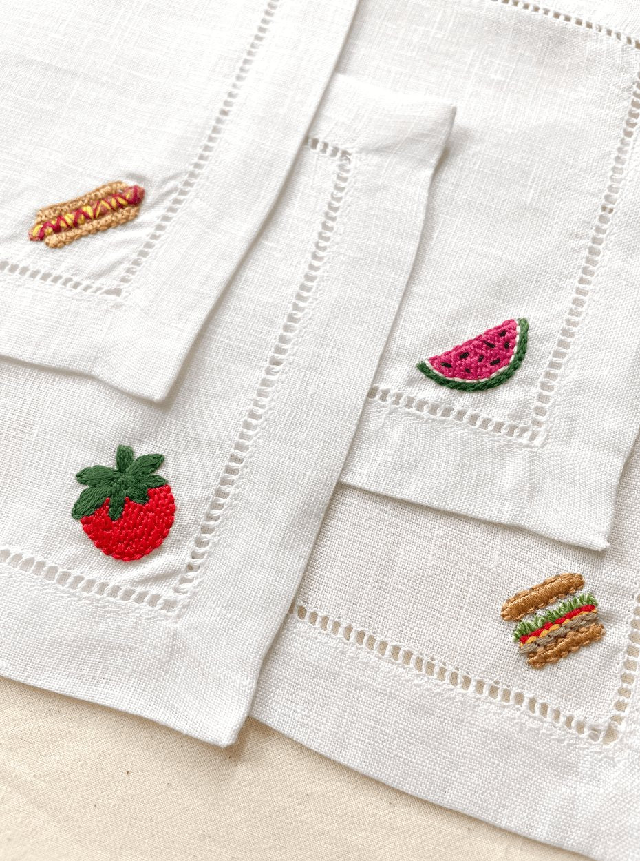 Set of 4 linen cocktail napkins featuring hand embroidered designs including a hotdog, tomato, watermelon, and hamburger on the corner of each napkin.