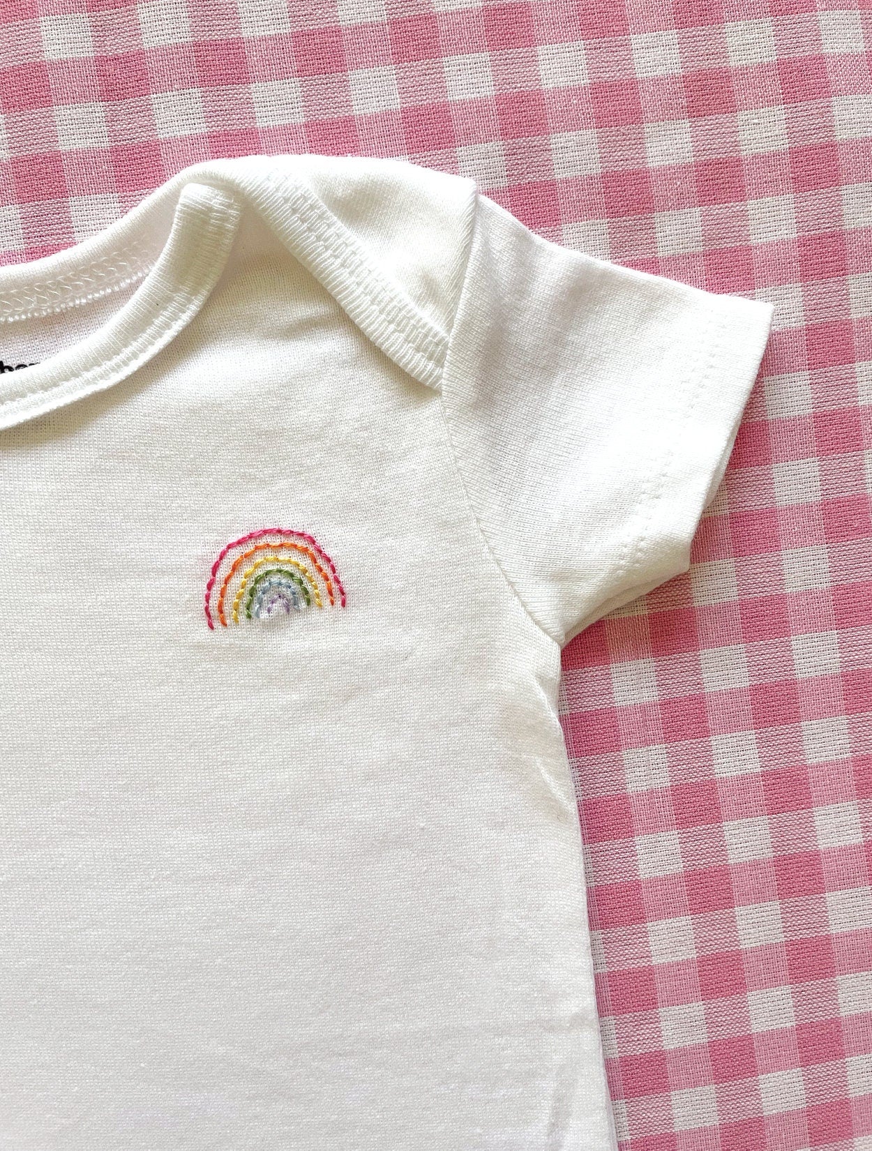 Close up image of hand embroidered onesie featuring rainbow design in bright colors on left chest