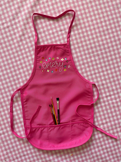 Hot pink children&#39;s apron embroidered with painted flowers around the name