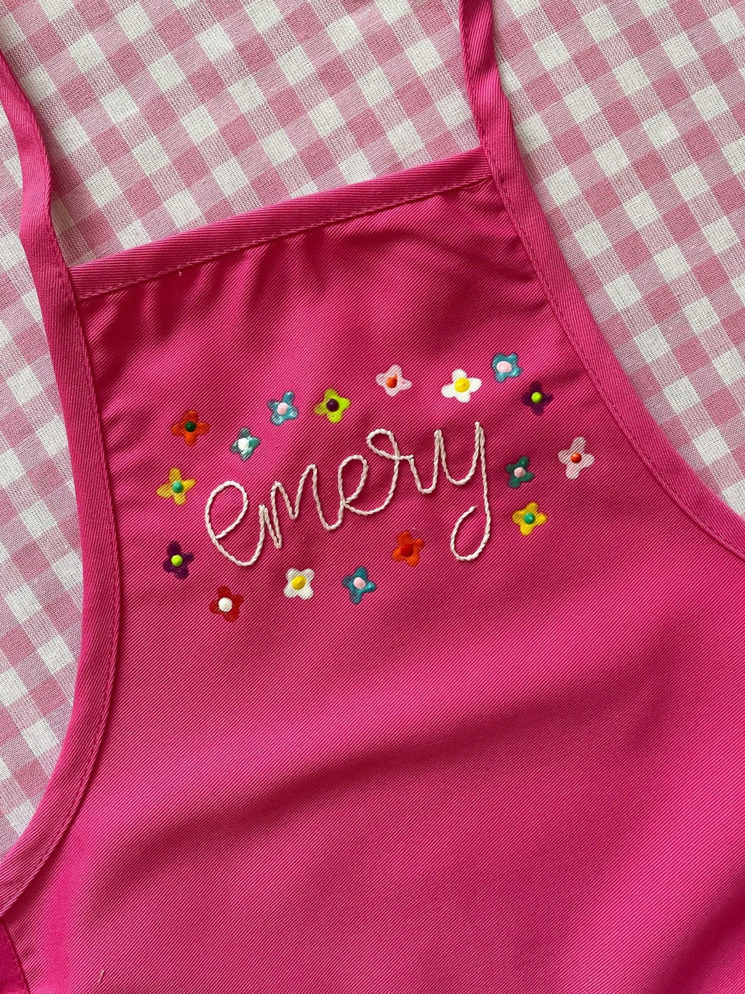 Top half of hot pink children&#39;s apron embroidered with painted flowers around the name